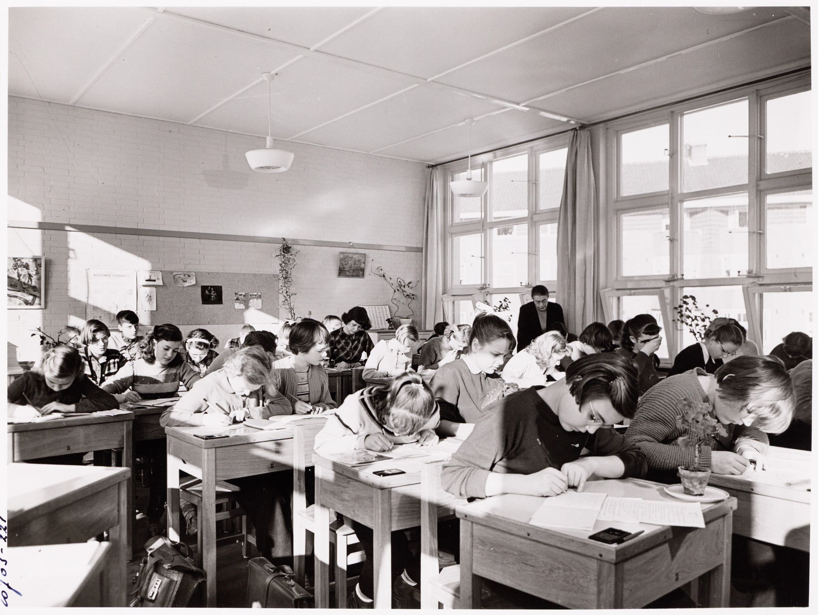 Meisjes krijgen les in een lokaal op de Aletta Jacobs-ULO, februari 1955. De meisjes zitten in een vol lokaal aan houten individuele schooltafeltjes en zijn zelfstandig aan het werk.